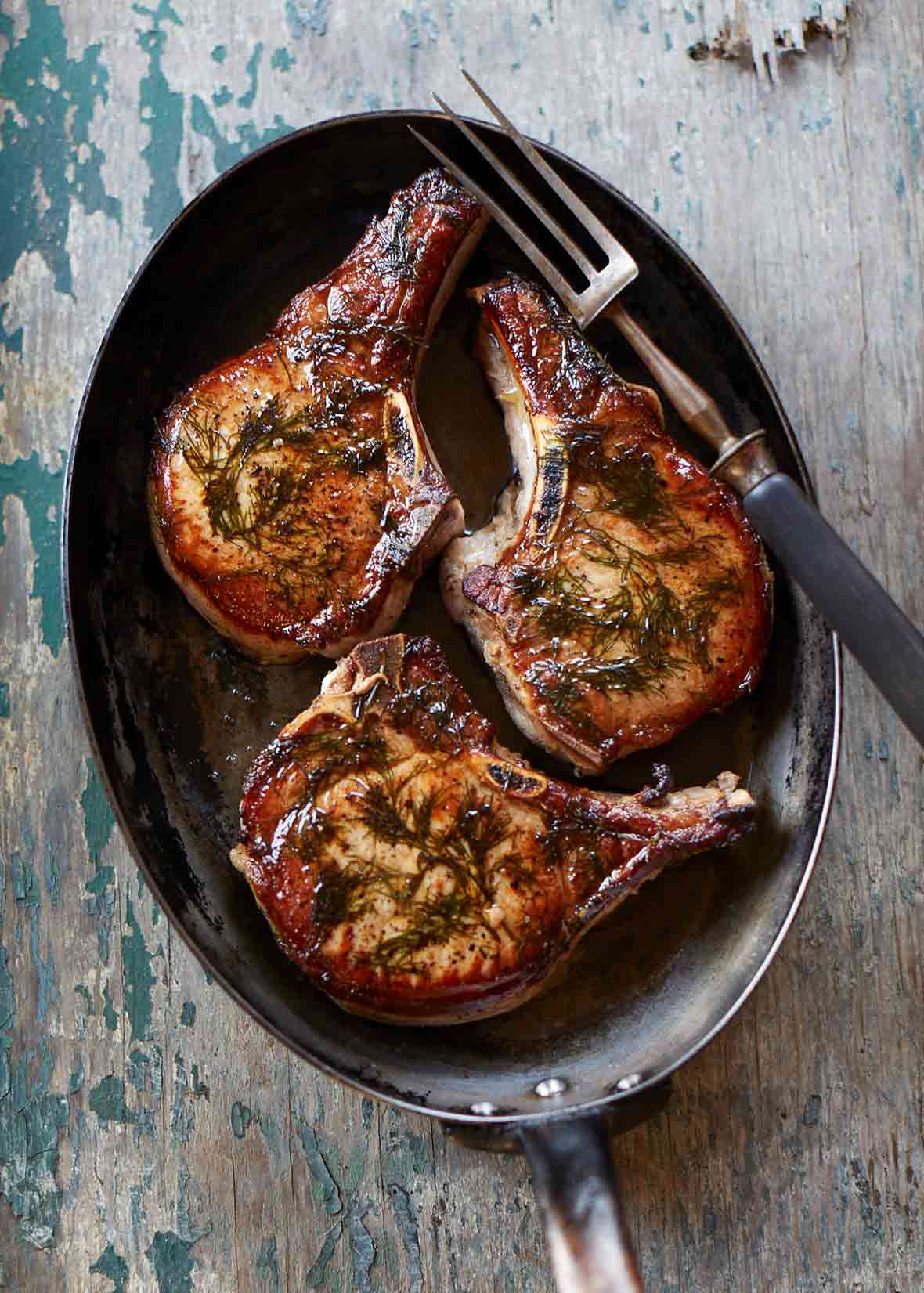Three seared pork chops in an oval skillet with a fork on top.