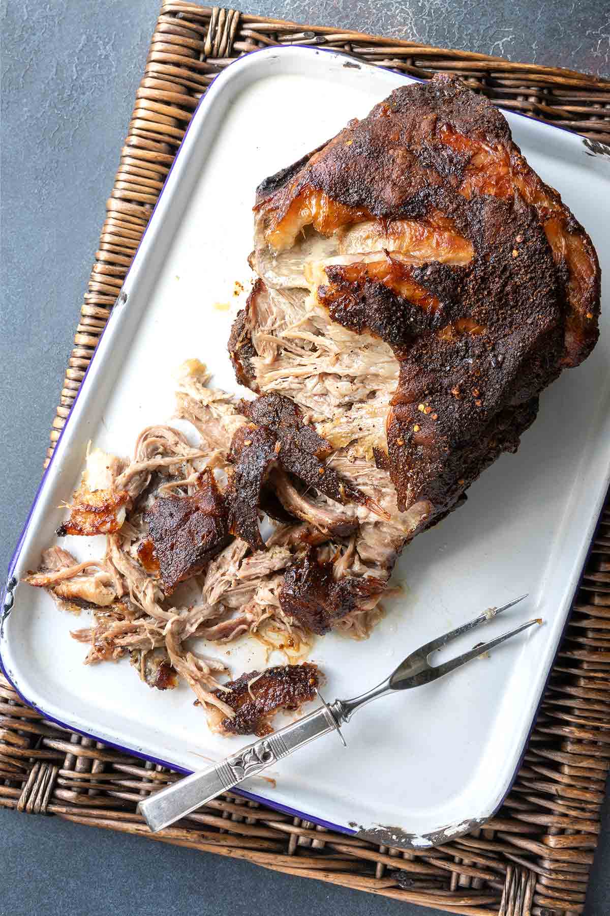 A partially shredded roast pork butt in a roasting pan with a fork nearby.