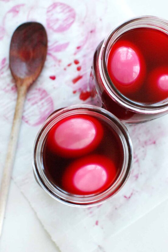Two jars filled with pickled eggs in a red liquid with a wooden spoon resting on the side.