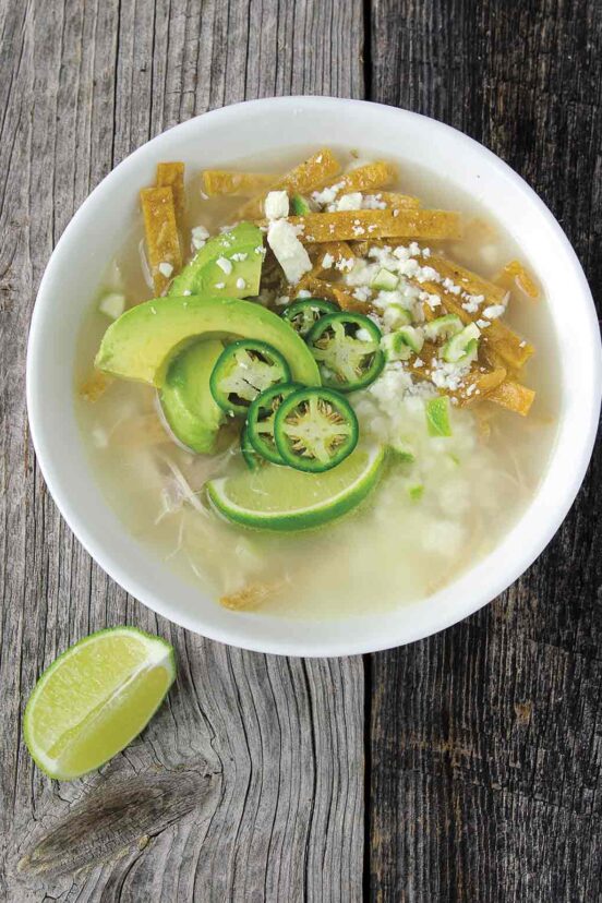 A white bowl filled with Mexican chicken soup, with fried tortilla strips, avocado slices, jalapeno slices, and cotija cheese, and a lime wedge on the side.