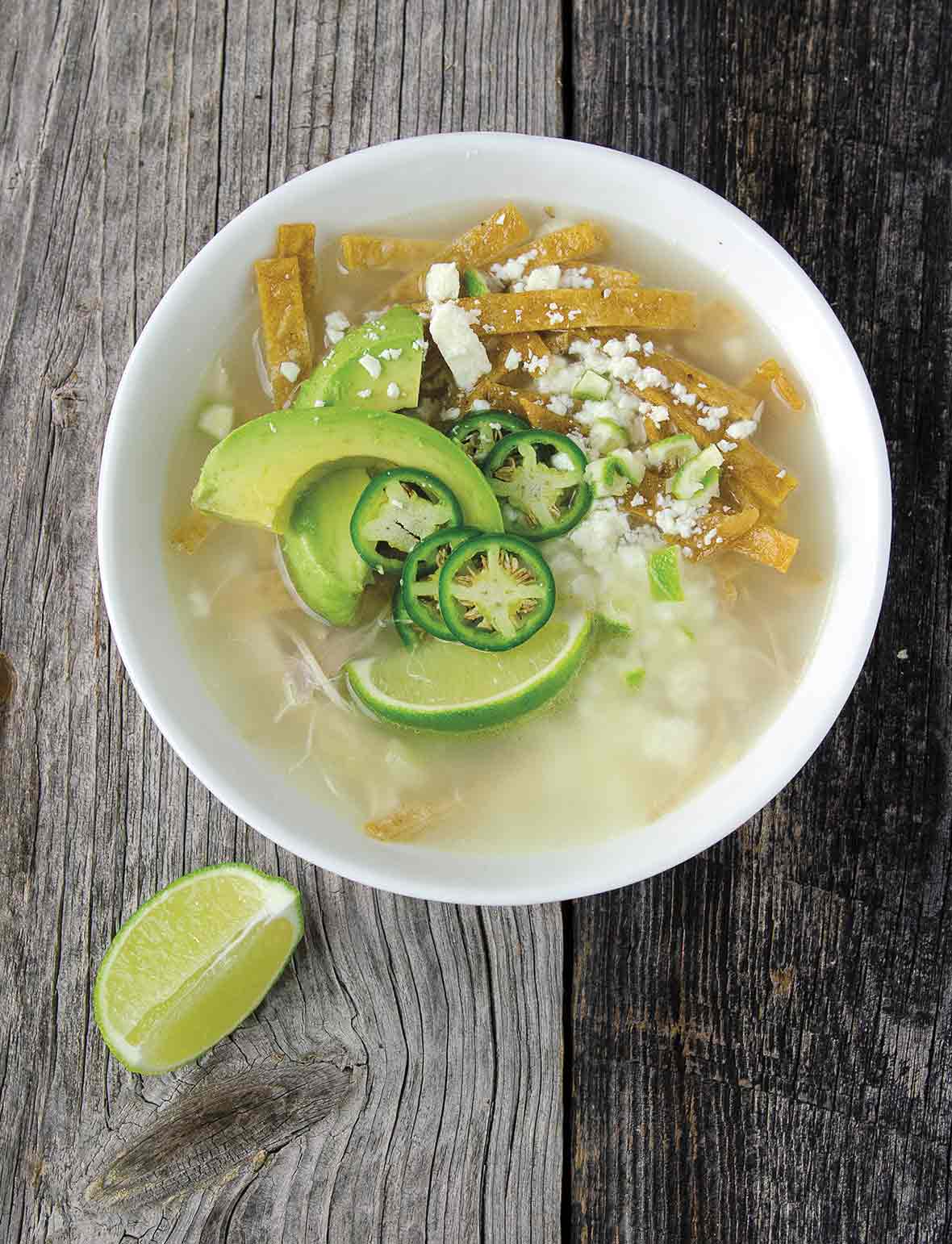 A white bowl filled with Mexican chicken soup, with fried tortilla strips, avocado slices, jalapeno slices, and cotija cheese, and a lime wedge on the side.