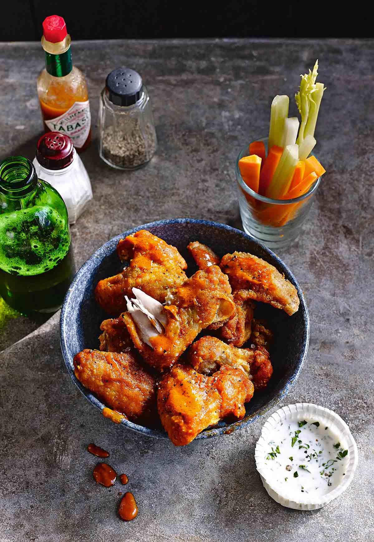 A bowls filled with buffalo chicken wings with a dish of ranch dip, a bottle of beer, and a glass of cut raw vegetables on the side.