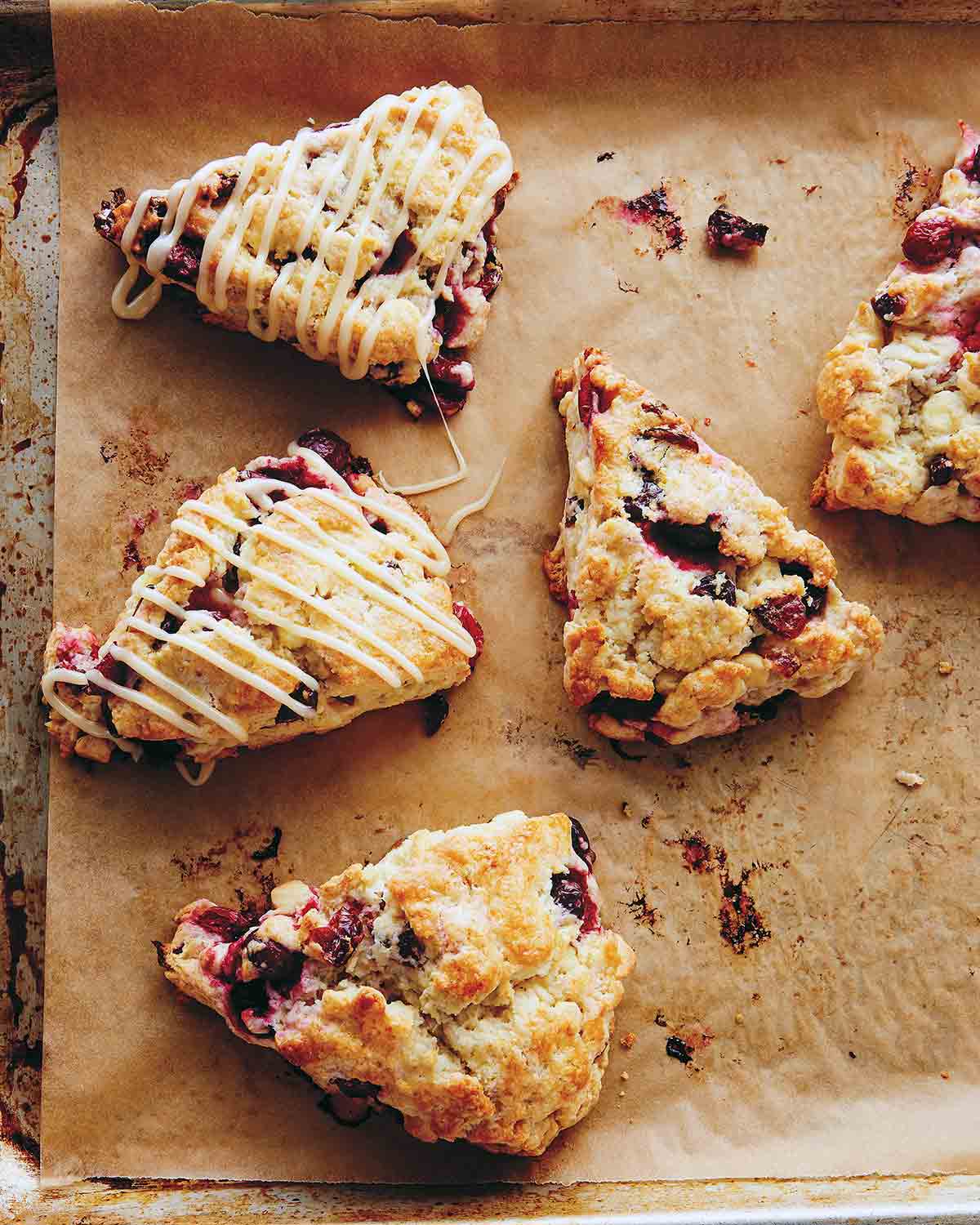 Five cranberry scones on a parchment-lined baking sheet and two are drizzled with white chocolate glaze.