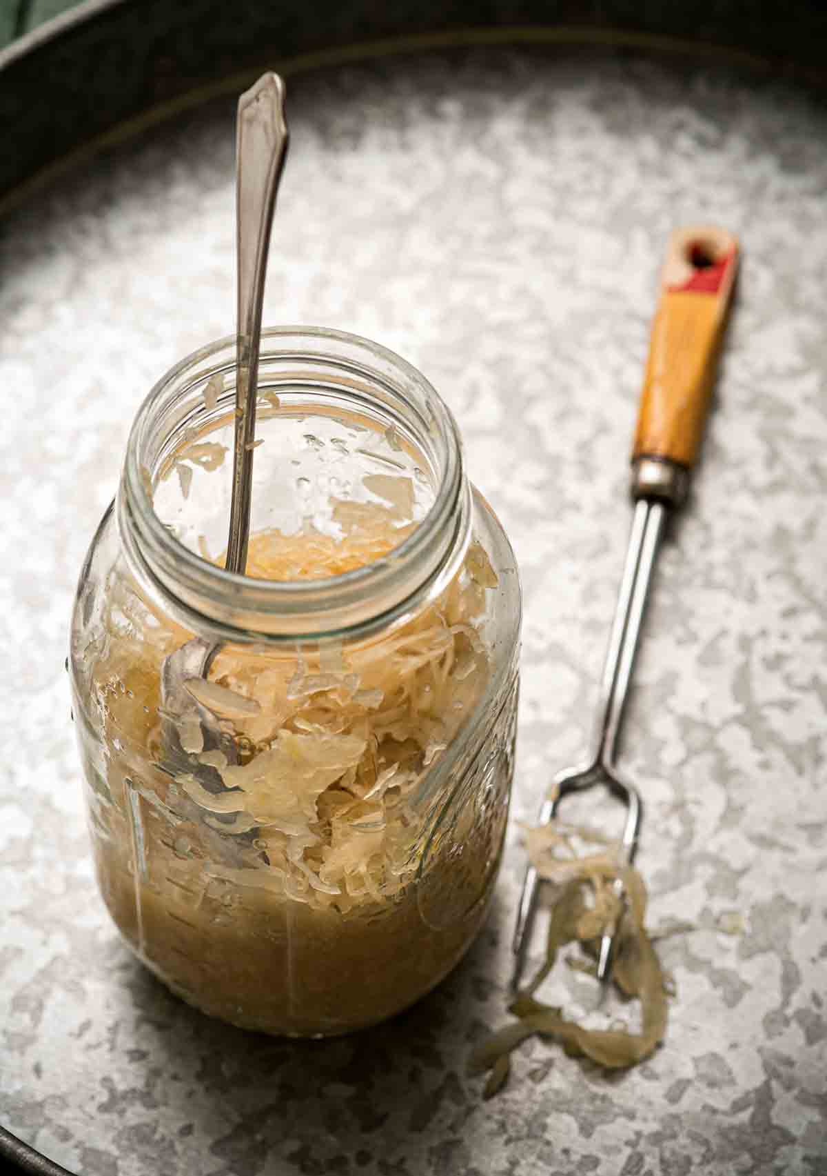 A jar of sauerkraut with a spoon standing up inside and a metal fork on the side.