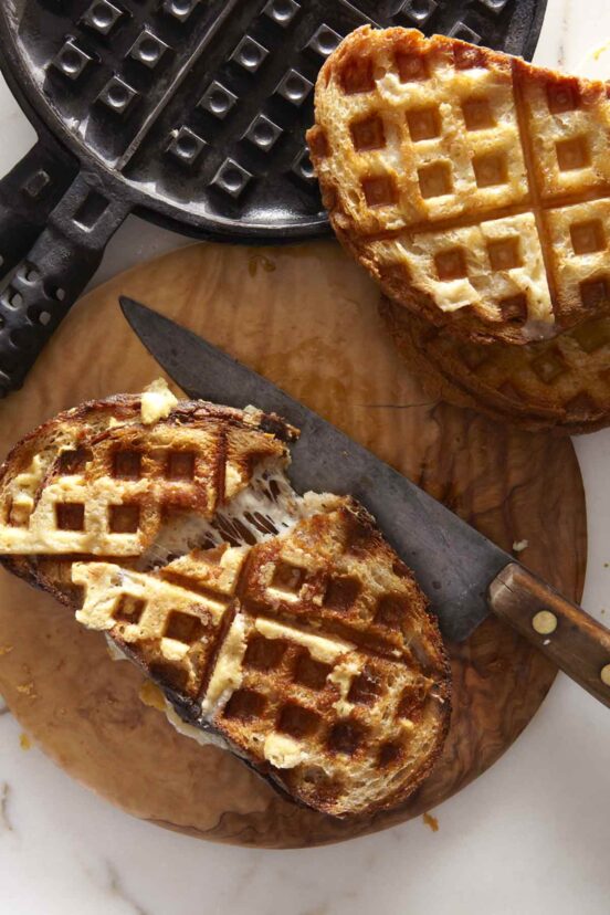 Two waffle iron grilled cheese sandwiches on a wooden board with a waffle maker to the side.