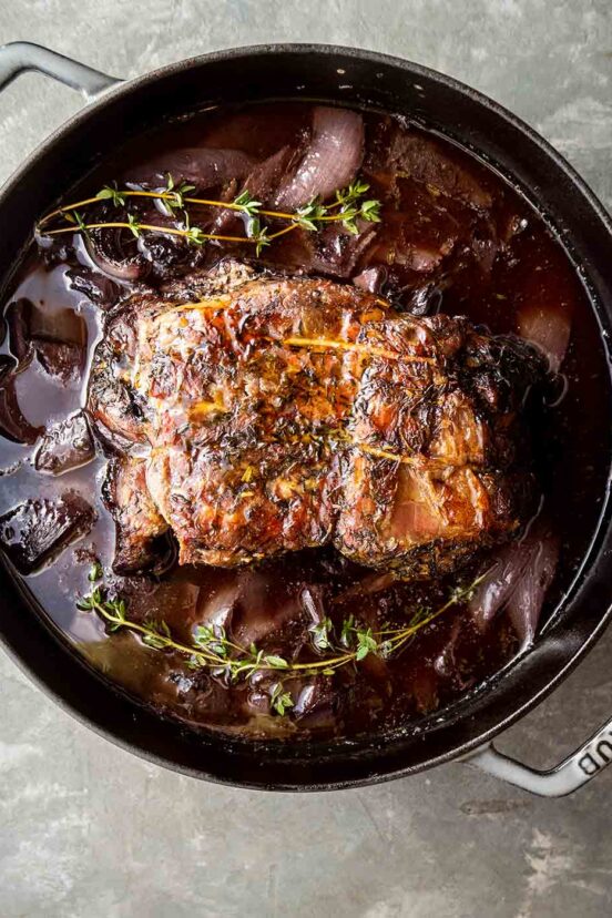 A braised pork shoulder in a Staub Dutch oven with red onions and fresh thyme in the liquid around the pork.