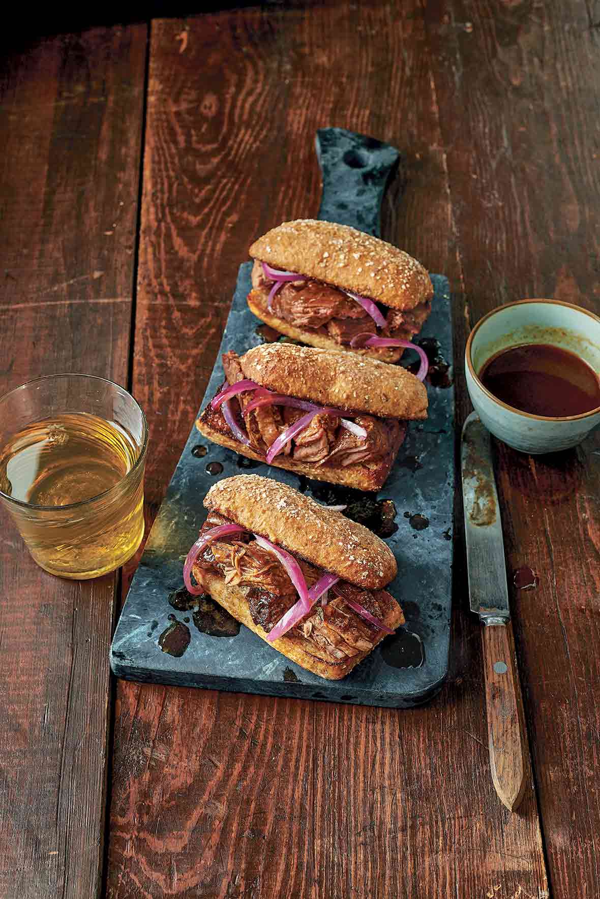 Three slow cooker French dip sandwiches on a slate board with a glass of beer, a knife, and bowl of sauce on the side.