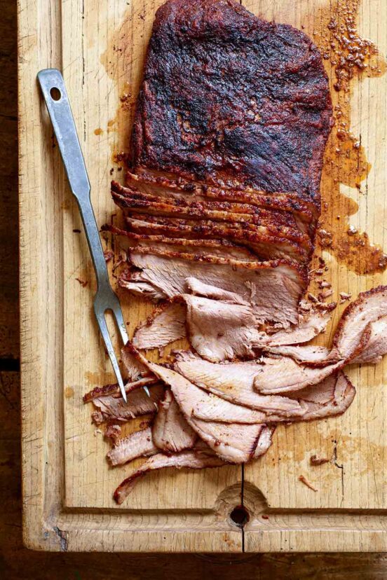 A partially sliced smoked brisket on a wooden carving board with a carving fork on the side.