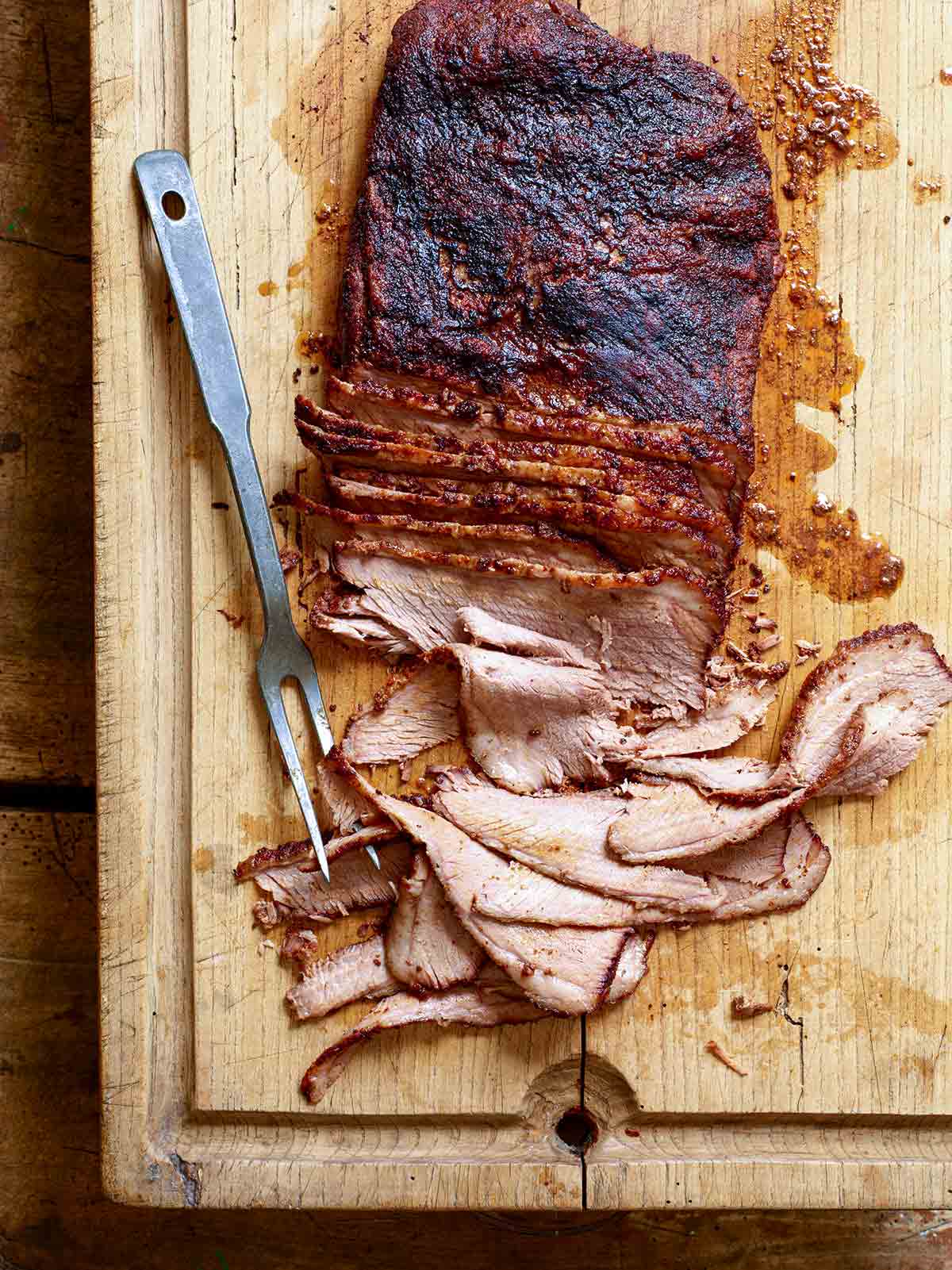 A partially sliced smoked brisket on a wooden carving board with a carving fork on the side.