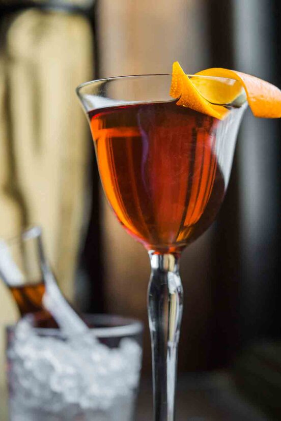 An aperitif glass filled with dark amber liquid and an orange twist on the rim.