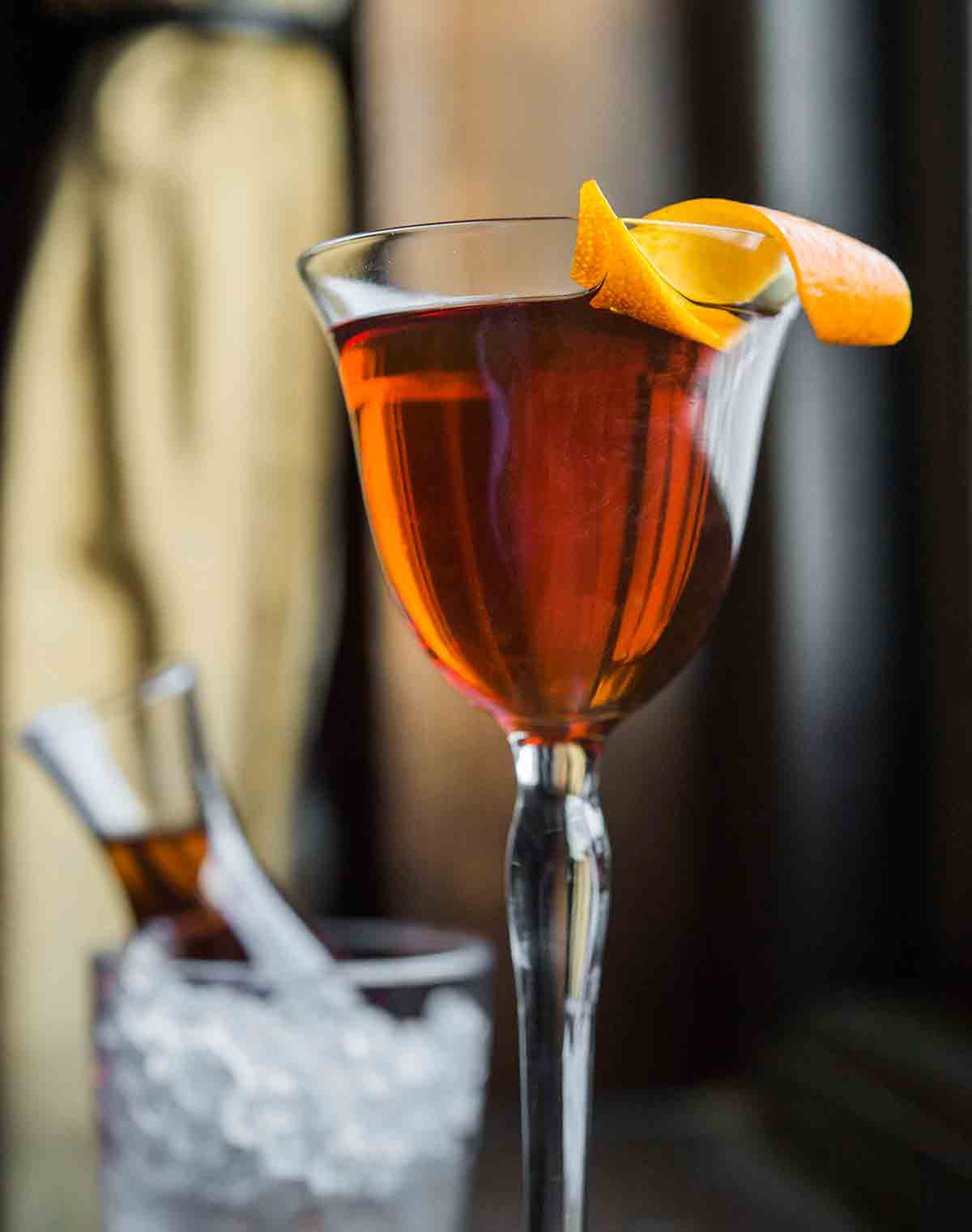 An aperitif glass filled with dark amber liquid and an orange twist on the rim.