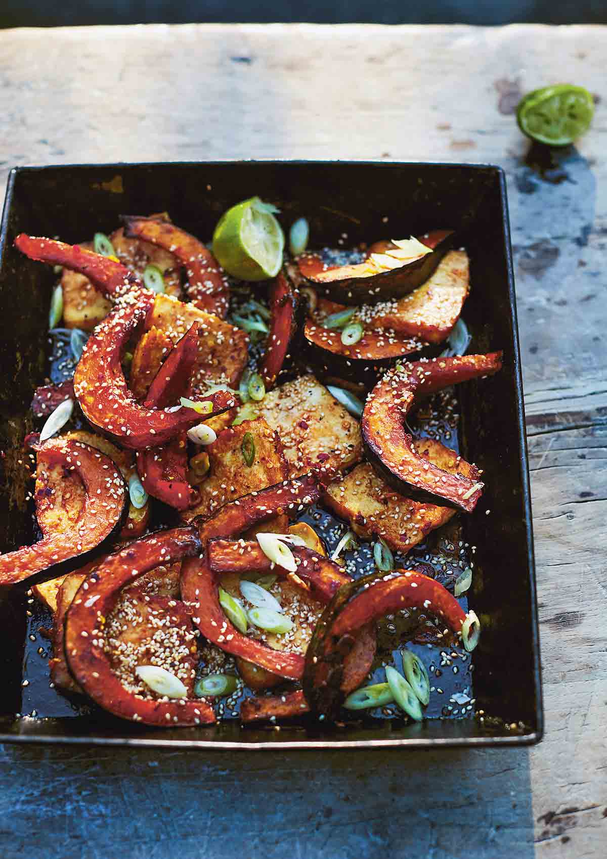 Wedges of roasted squash in a black square serving dish, scattered with sesame seeds and lime halves on the side.