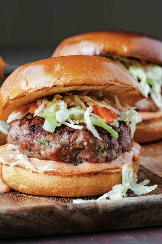 Three grilled pork burgers topped with coleslaw and yum yum sauce on a wooden serving tray.