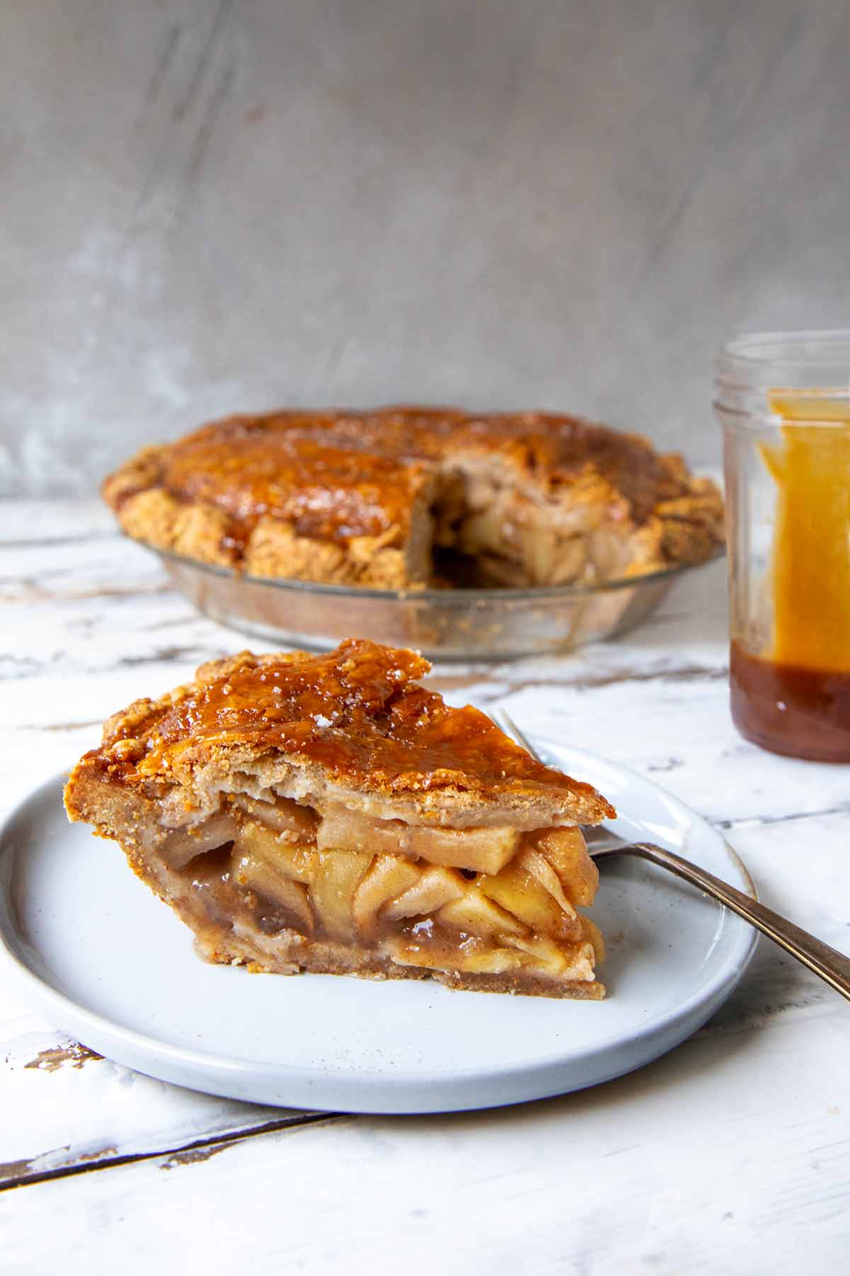 A slice of caramel apple pie on a white plate with the remaining pie and a jar of caramel in the background.