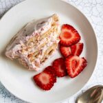 A serving of strawberry icebox cake on a plate with sliced strawberries and a spoon on the side.