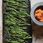 A cast-iron griddle filled with cooked broccolini and a bowl of red paste on the side.