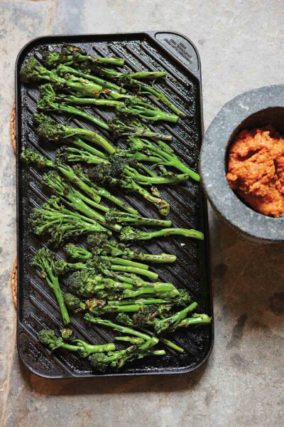 A cast-iron griddle filled with cooked broccolini and a bowl of red paste on the side.