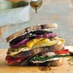 A grilled vegetable and goat cheese sandwich, made with grilled onion, eggplant, and squash on a cutting board being drizzled with balsamic vinaigrette.