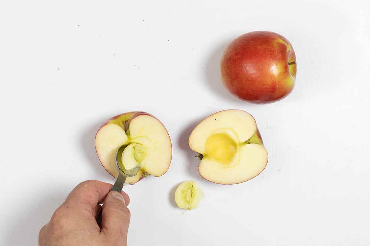 A whole apple and a halved apple with a person using a spoon to remove the core from the center of the halved apple.
