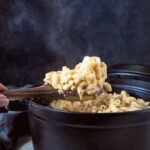A hand using a wooden spoon to lift a scoop of macaroni and cheese with zucchini out of a cast iron pot.