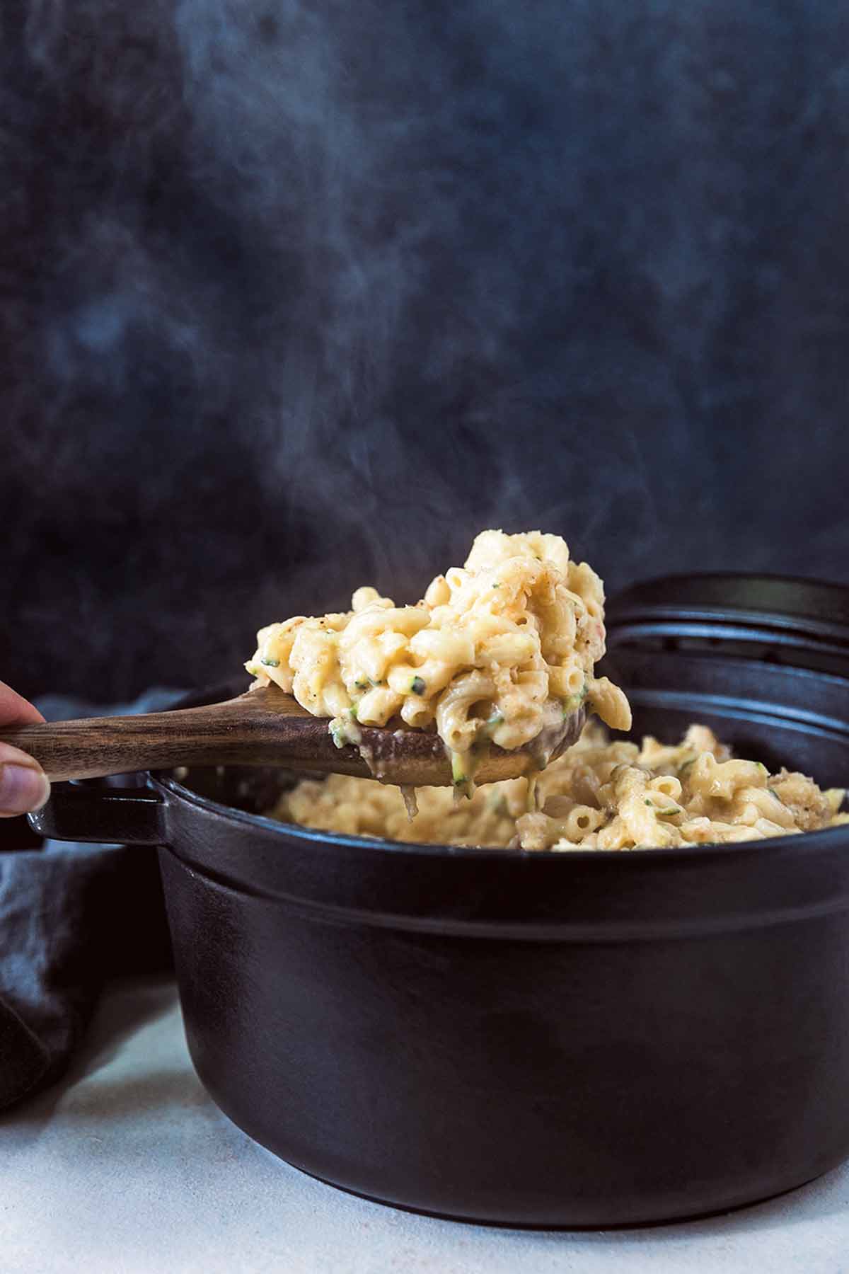A hand using a wooden spoon to lift a scoop of macaroni and cheese with zucchini out of a cast iron pot.