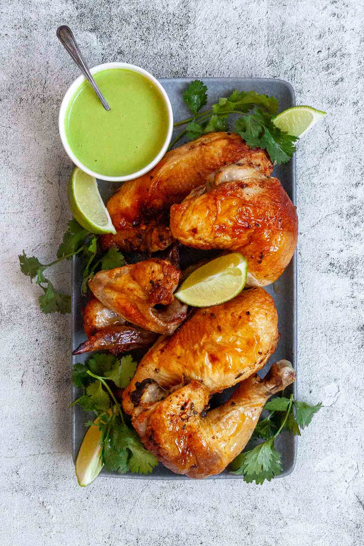 A carved Peruvian chicken on a rectangular platter with lime wedges, cilantro, and a dish of green sauce on the side.