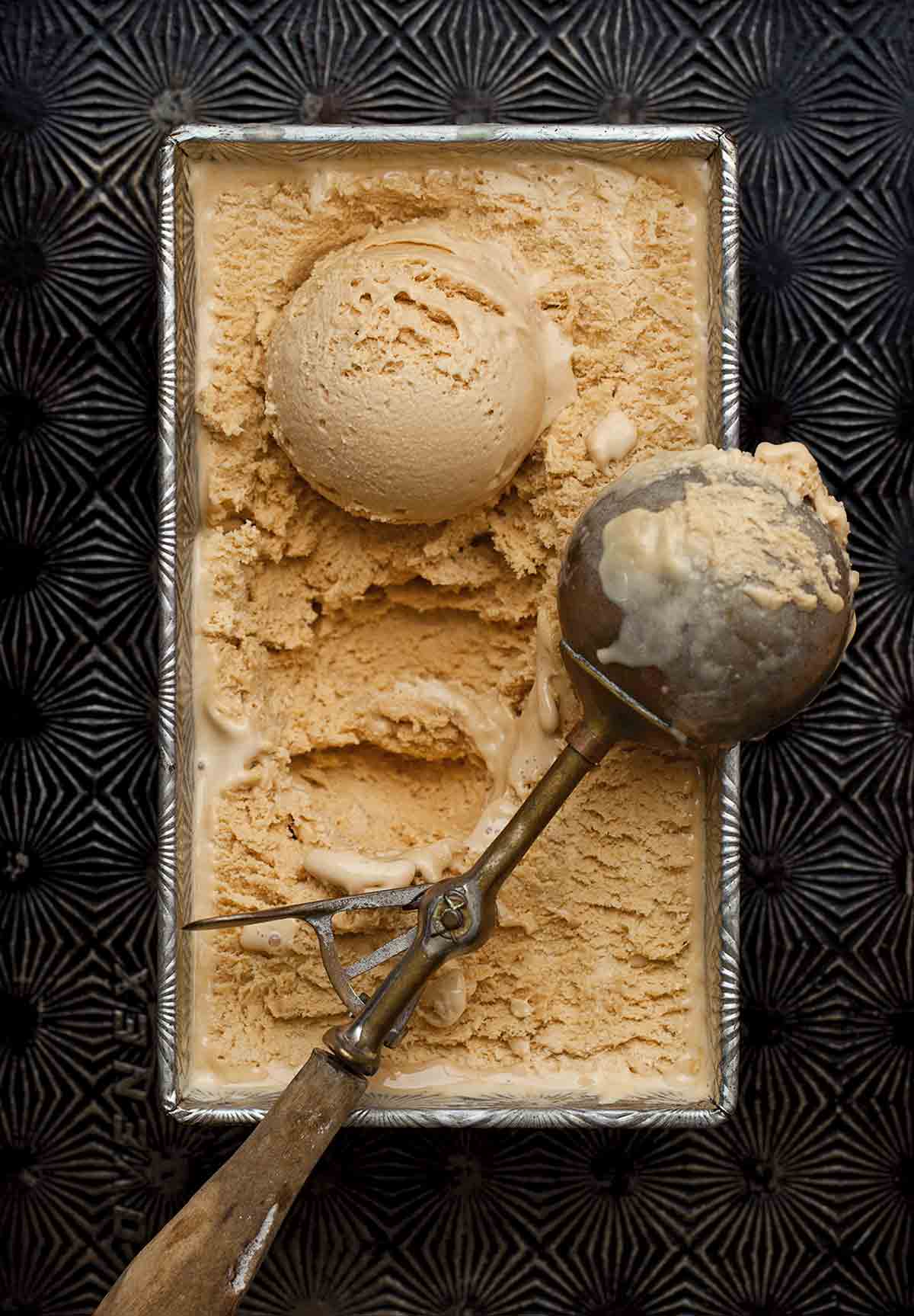 Salted caramel ice cream in a loaf pan on a pattern baking sheet with a scoop and scooper on top.