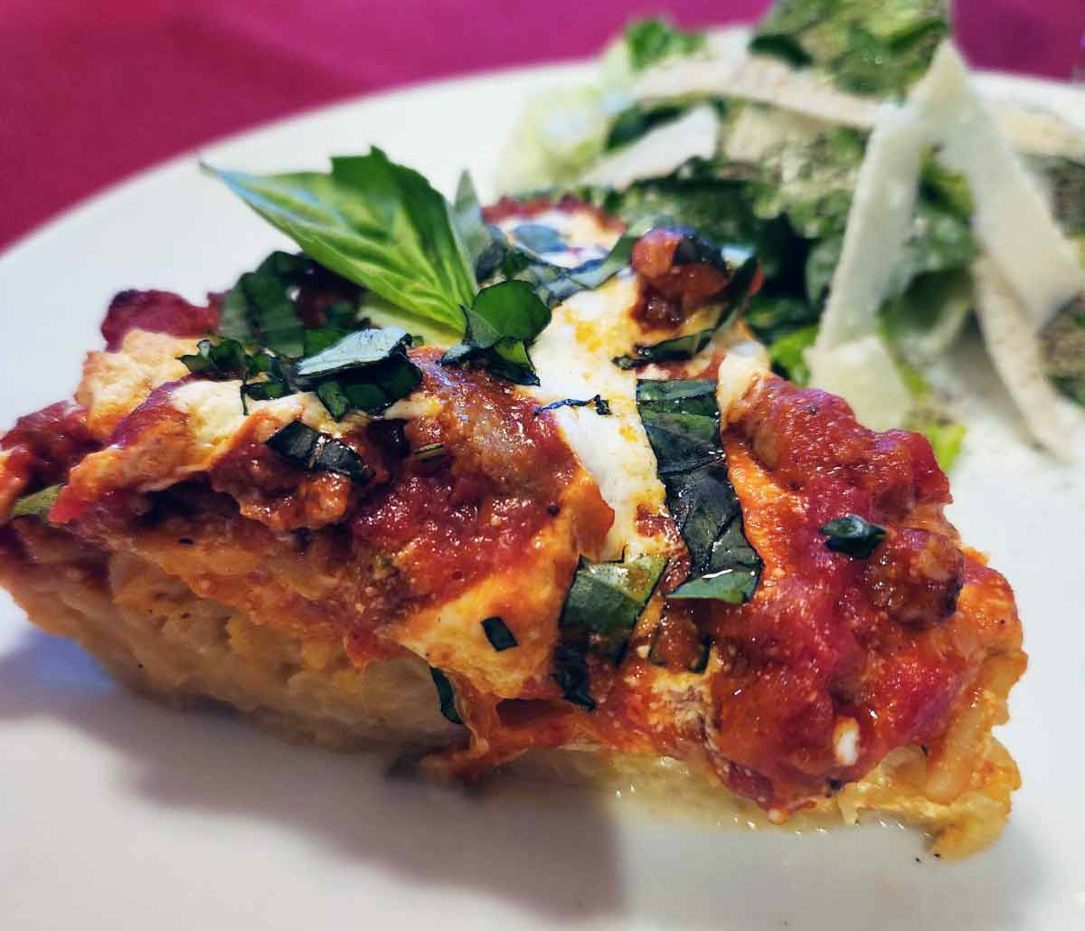 A slice of spaghetti pie on a white plate with some salad topped with shaved Parmesan in the background.