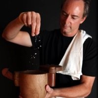 A man in a black shirt sprinkling salt into a wooden bowl