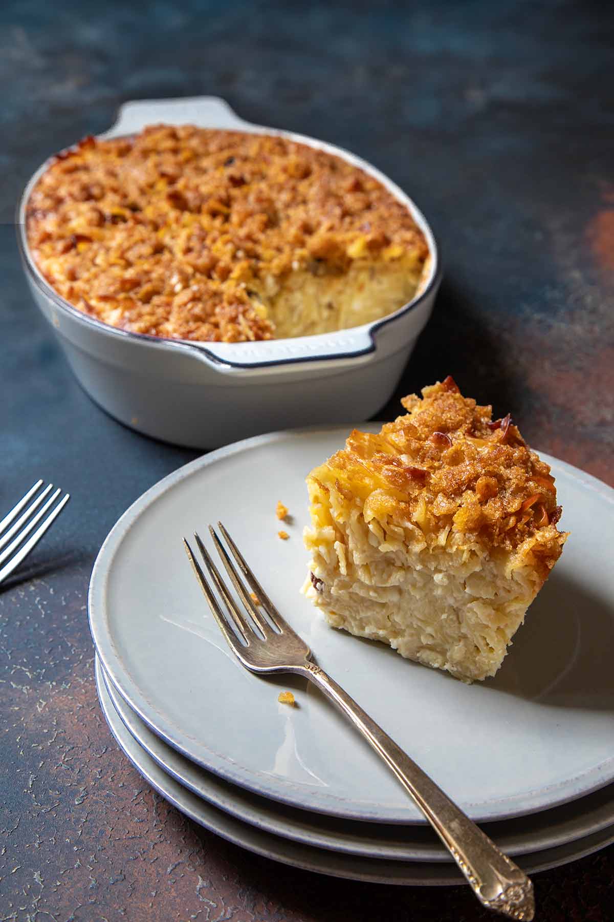 A casserole dish of sweet noodle kugel; in front is a plate with a slice of kugel on it.