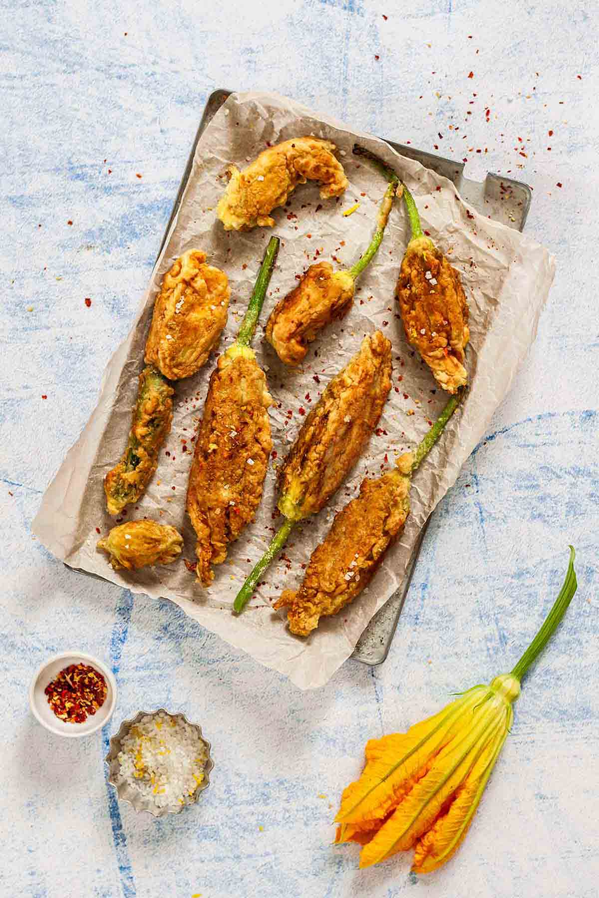 A large pan lined with parchment paper and 8 deep fried zucchini flowers, sprinkled with lemon zest, salt, and chile flakes.