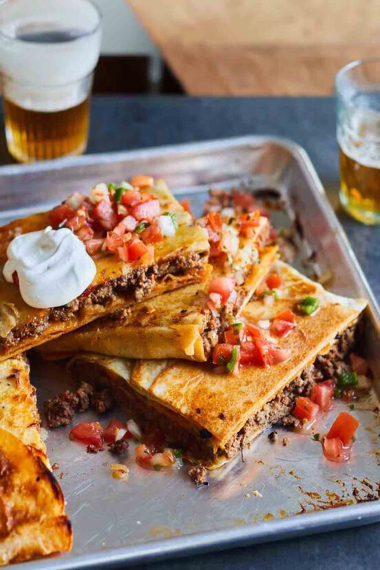 A cut sheet pan taco bake in a rimmed sheet pan topped with pico de gallo and sour cream.
