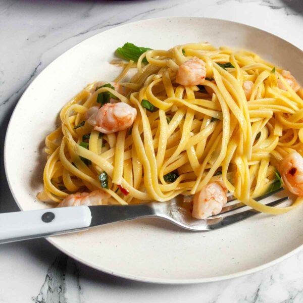 A plate of shrimp linguine with a fork on the side and a skillet of the pasta in the background.