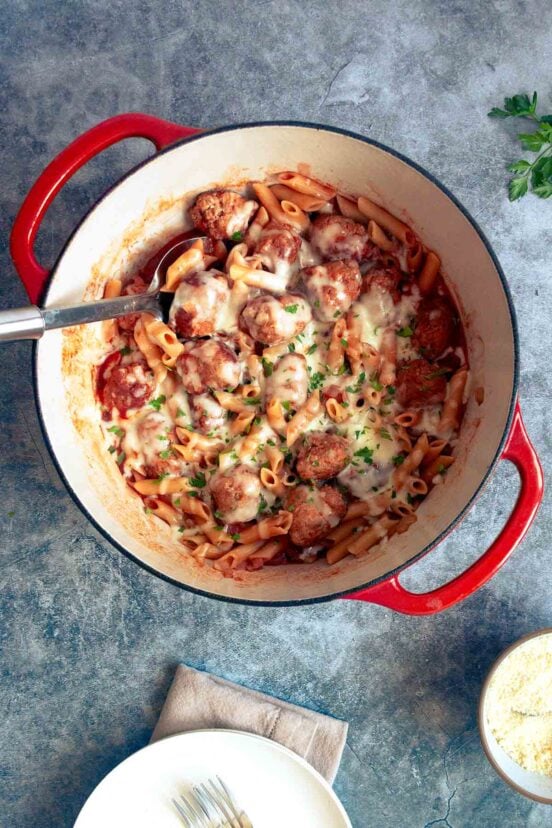 A red Dutch oven filled with pasta and meatballs, covered in cheese.