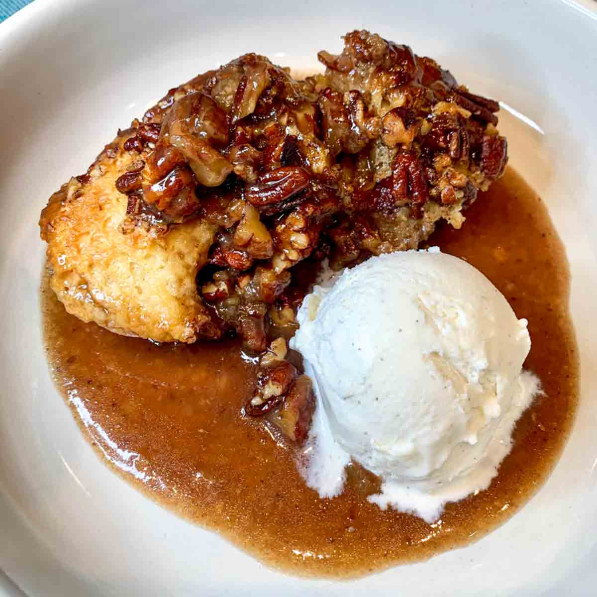 A serving of pecan pie cobbler in a white bowl with a scoop of vanilla ice cream next to it.