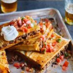 A cut sheet pan taco bake in a rimmed sheet pan topped with pico de gallo and sour cream.