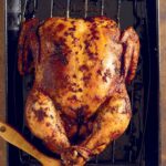 A whole spicy roast chicken on a rack set inside a baking dish with a brush lying beside the chicken.