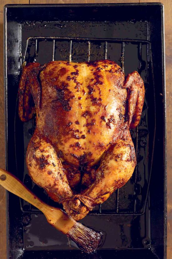 A whole spicy roast chicken on a rack set inside a baking dish with a brush lying beside the chicken.