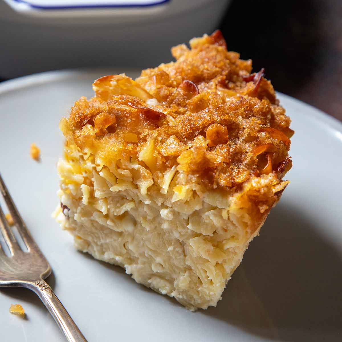 A casserole dish of sweet noodle kugel; in front is a plate with a slice of kugel on it.