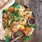 Crispy chicken with lemon and capers in a large serving tray, with a bowl of capers and a bowl of peppercorns. The chicken is garnished with lemon slices and parsley.