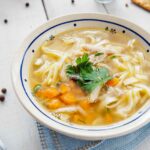 A bowl of chicken soup with carrots and noodles on a white table.
