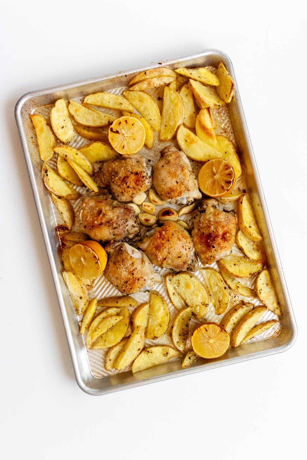 A sheet pan filled with roasted potato wedges, lemon halves and crispy chicken thighs.
