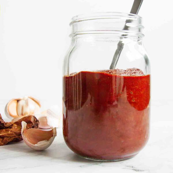 A jar of homemade adobo sauce with a spoon resting inside and garlic cloves and dried chipotles on the side.