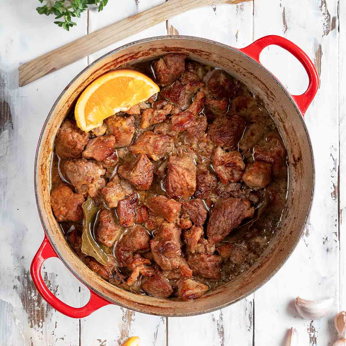 A red Dutch oven filled with braised pork cubes with an orange wedge on top and a few orange wedges, garlic cloves, oregano, and spoon on the side.