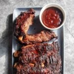 A rack of barbecued beef back ribs on a rectangular platter with a dish of barbecue sauce on the side.
