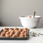 A tray of unbaked bbq meatballs with a set of measuring spoons beside it and a bowl of barbecue sauce in the background.