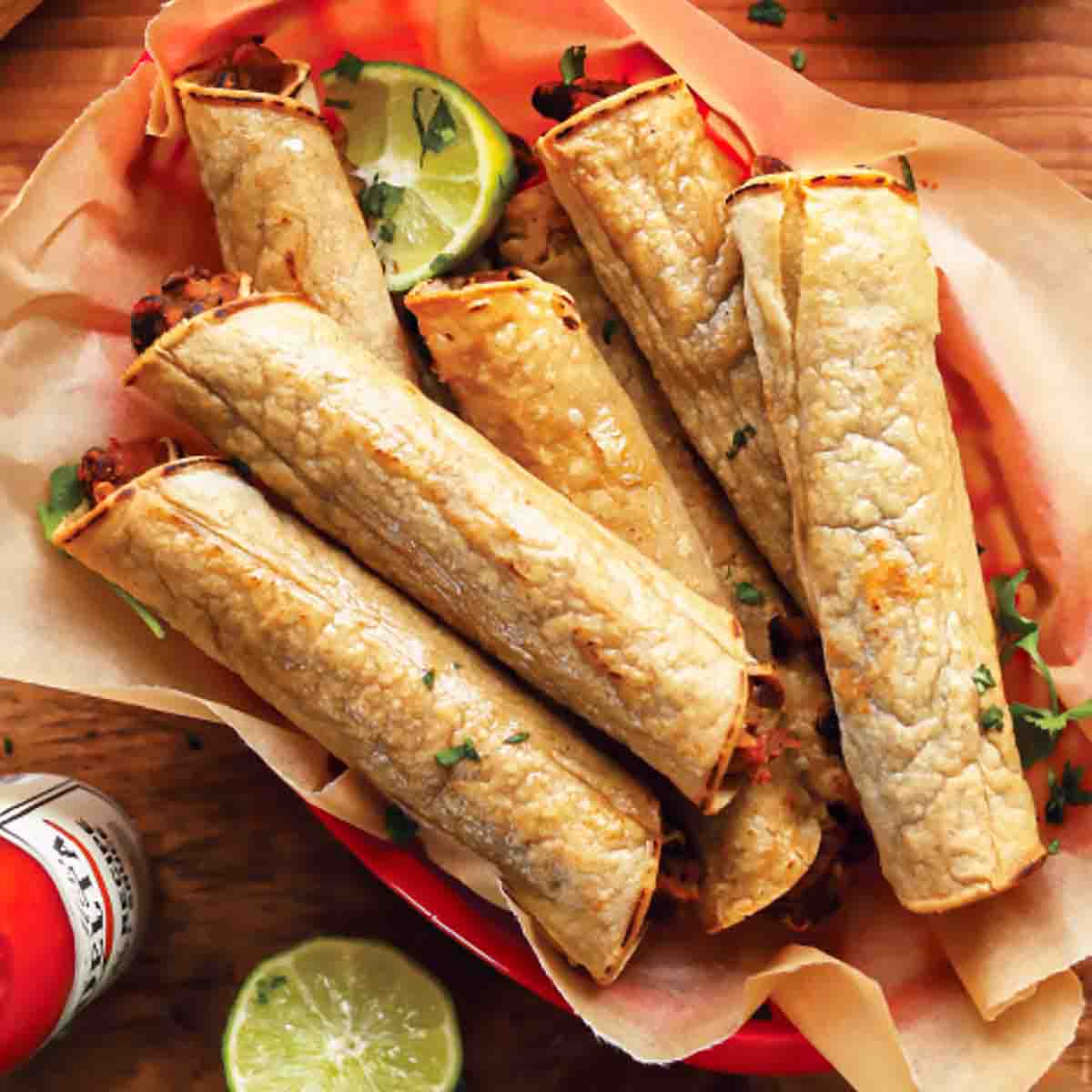A basket of black bean taquitos with lime halves and hot sauce on the side.