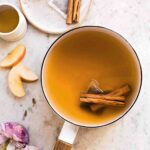 A white enamel pot filled with chai spiced apple cider and two cinnamon sticks.
