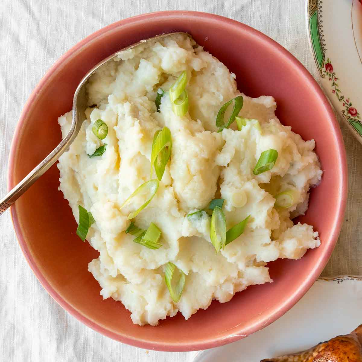 A bowl of cheesy mashed potatoes, topped with scallions on a table with turkey and cranberry sauce.