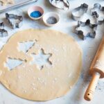 Christmas sugar cookies being rolled out and placed on a sheet pan, with cookie cutters, a rolling pin, sprinkles, and flour nearby.